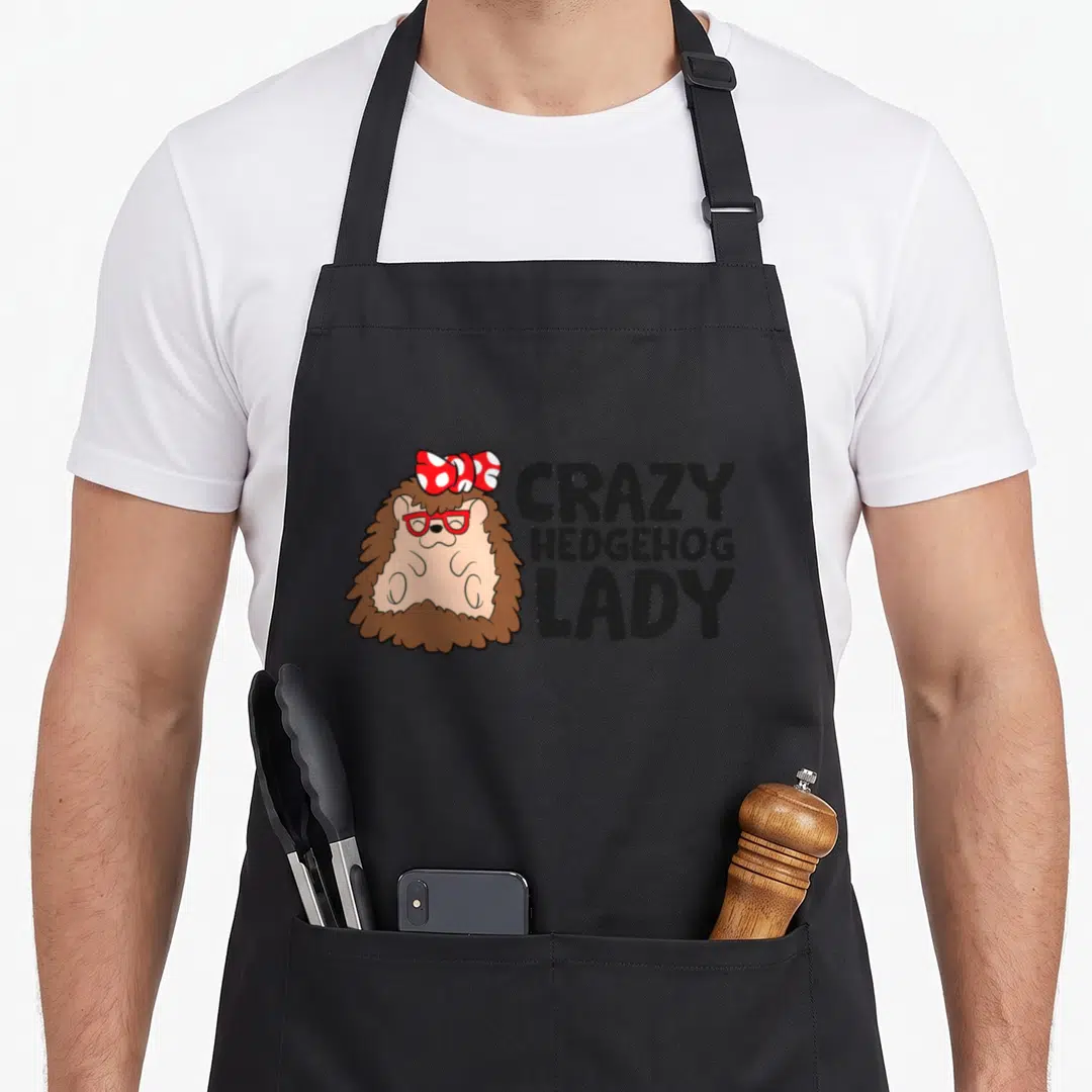 Comfort crazy hedgehog lady apron - Cartoon brown hedgehog with red glasses and polka dot bow next to stacked black text — shown on a black cotton canvas apron against a white background
