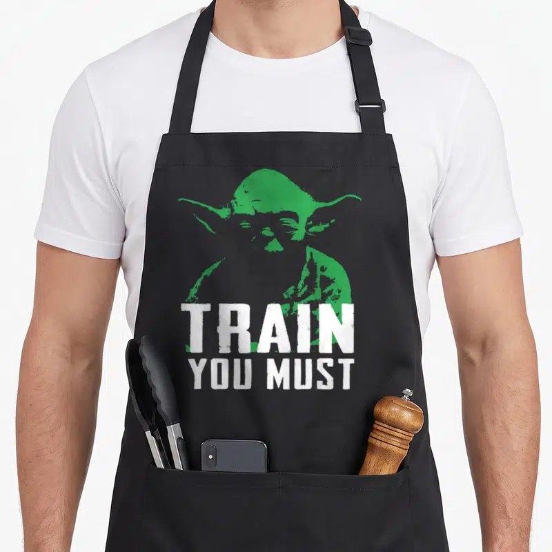 Train yoda apron - Green Yoda silhouette with white distressed text reading TRAIN YOU MUST — shown on a black cotton canvas apron against a white background