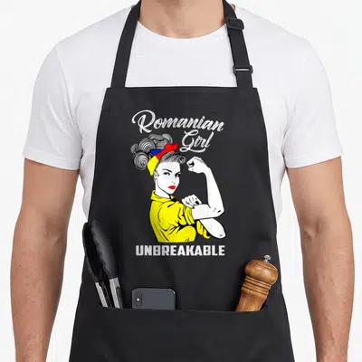 Unbreakable romanian apron - Cartoon woman flexing arm with Romanian flag headband and text Romanian Girl UNBREAKABLE — shown on a black cotton canvas apron against a white background