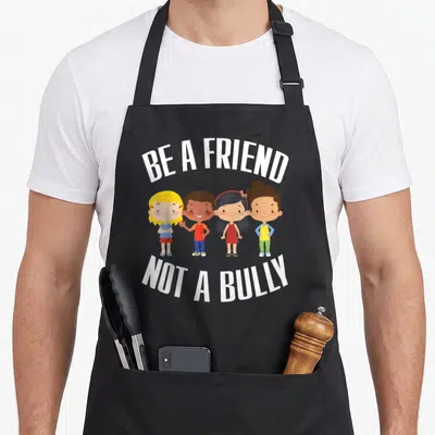 Anti best friend aprons - Four cartoon children standing in a row with text BE A FRIEND NOT A BULLY in white, blue — shown on a black cotton canvas apron against a white background