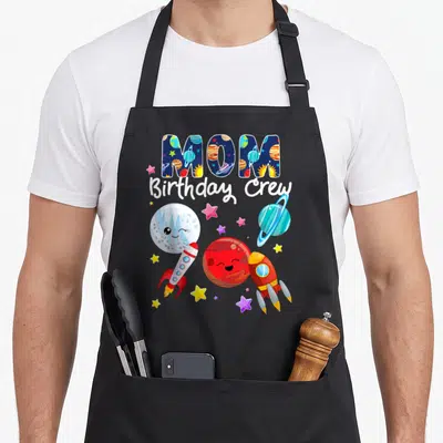 Crew outer birthday apron - MOM Birthday Crew text with cartoon planets rockets and stars in blue red yellow and white — shown on a black cotton canvas apron against a white background