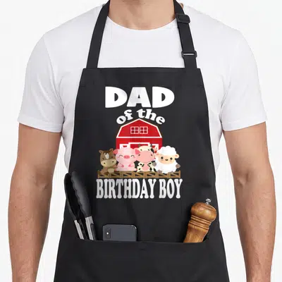 Animal farming apron - DAD of the BIRTHDAY BOY text above red barn with four cartoon animals behind brown fence — shown on a black cotton canvas apron against a white background