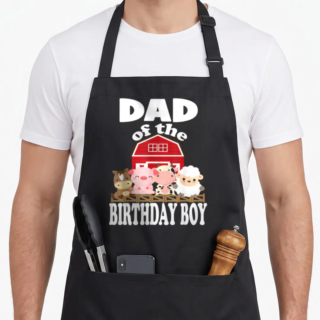 Animal farming apron - DAD of the BIRTHDAY BOY text above red barn with four cartoon animals behind brown fence — shown on a black cotton canvas apron against a white background