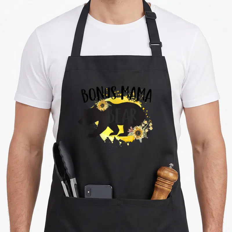 Sunflower mama bear apron - Black bear silhouette with sunflowers and text BONUS MAMA BEAR on yellow splatter — shown on a black cotton canvas apron against a white background