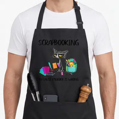 Scrapbooking cat apron - Grey cartoon cat with teal backpack and pink paper under text Because Murder Is Wrong — shown on a black cotton canvas apron against a white background