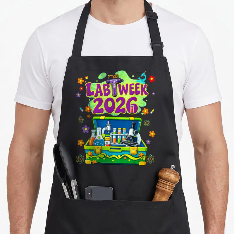 Week laboratory apron - Lab Week 2026 purple text over green splash with open toolbox containing microscopes, flasks — shown on a black cotton canvas apron against a white background