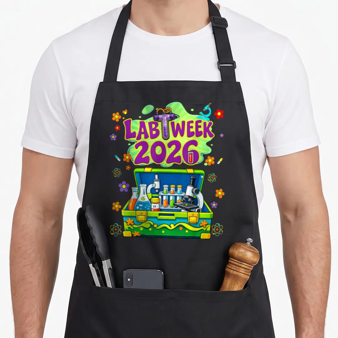 Week laboratory apron - Lab Week 2026 purple text over green splash with open toolbox containing microscopes, flasks — shown on a black cotton canvas apron against a white background