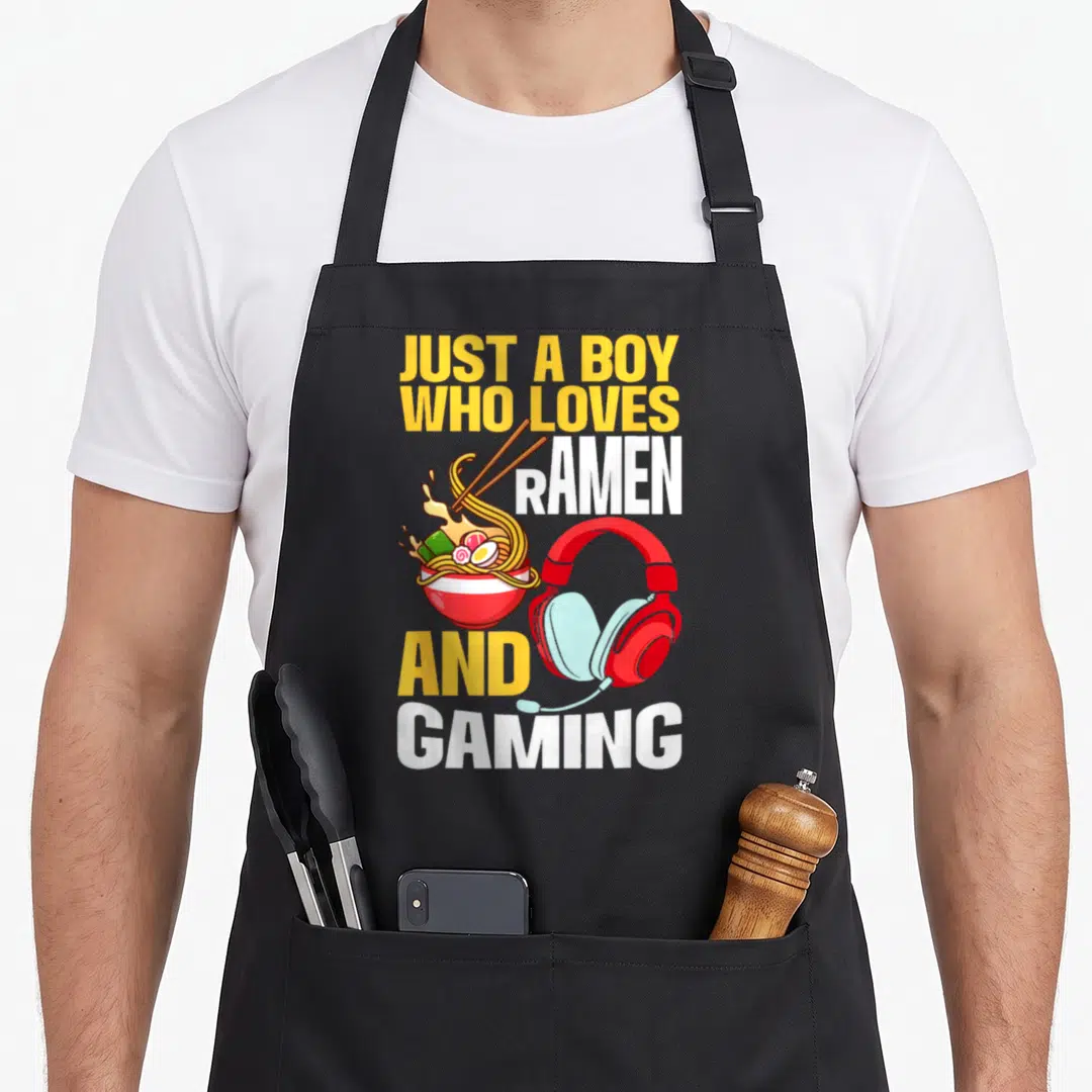 Ramen video game apron - Cartoon ramen bowl with chopsticks and red gaming headphones under stacked yellow and white — shown on a black cotton canvas apron against a white background