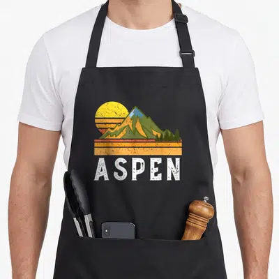 Vacation colorado apron - Retro mountain graphic with yellow sun, green peaks, pine trees, orange stripes, and white — shown on a black cotton canvas apron against a white background