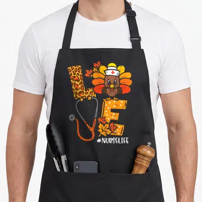 Nurse thanksgiving apron - Cartoon turkey in nurse hat on orange E with stethoscope O and #NURSELIFE text — shown on a black cotton canvas apron against a white background
