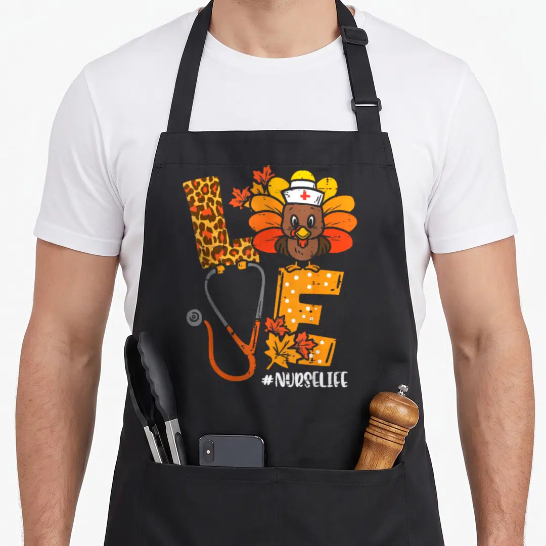 Nurse thanksgiving apron - Cartoon turkey in nurse hat on orange E with stethoscope O and #NURSELIFE text — shown on a black cotton canvas apron against a white background