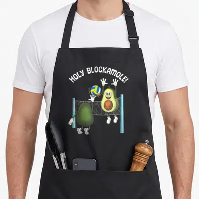Holy guacamole apron - Two cartoon avocados playing volleyball under holy blockamole text — shown on a black cotton canvas apron against a white background