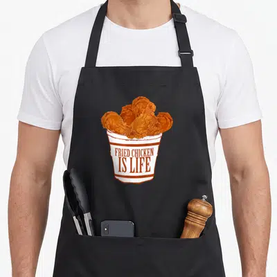 Fried bucket chicken apron - Cartoon bucket with golden brown fried chicken and brown text FRIED CHICKEN IS LIFE — shown on a black cotton canvas apron against a white background