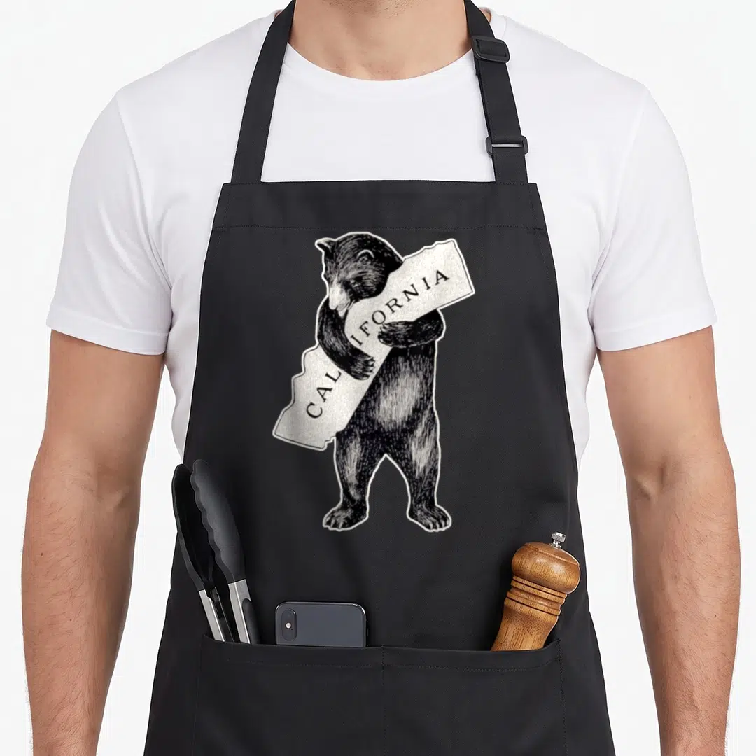 Cali bear apron - Black bear standing and hugging a white irregular sign printed with CALIFORNIA — shown on a black cotton canvas apron against a white background