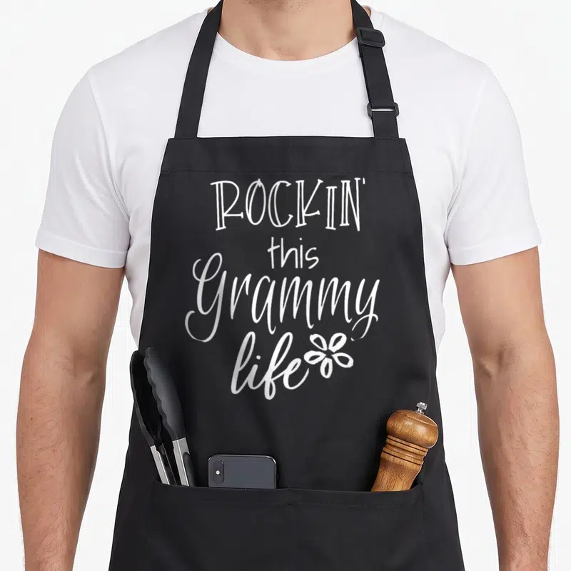 Rockin grammy apron - White stacked text reading ROCKIN this Grammy life next to a small flower outline — shown on a black cotton canvas apron against a white background