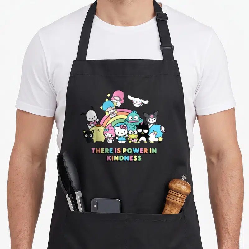 Power friends apron - Sanrio characters clustered around a rainbow with colorful text reading THERE IS POWER IN KINDNESS — shown on a black cotton canvas apron against a white background
