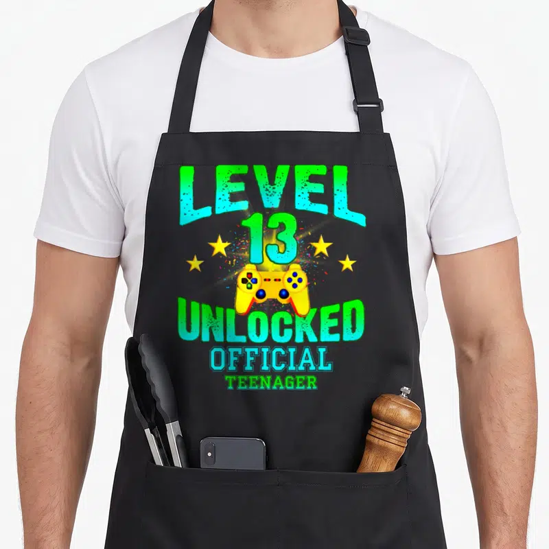Cute level birthday apron - Vertical text reading LEVEL 13 UNLOCKED OFFICIAL TEENAGER surrounding a yellow game — shown on a black cotton canvas apron against a white background