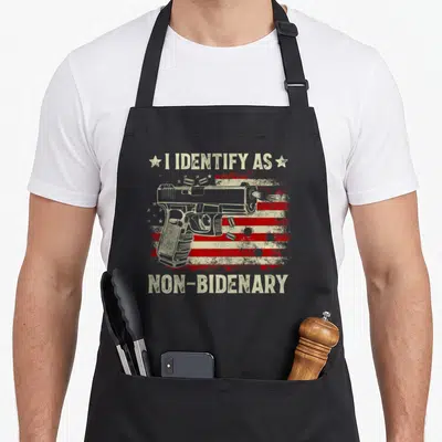 Identify american flag apron - Grey handgun over distressed red beige flag with text I Identify As Non-Bidenary flanked — shown on a black cotton canvas apron against a white background
