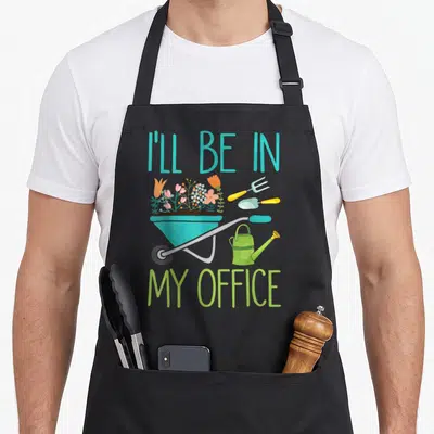 Ill spring aprons - Teal wheelbarrow with flowers and tools under text I'll Be In My Office — shown on a black cotton canvas apron against a white background