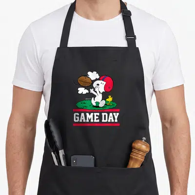 Woodstock football apron - Snoopy in red helmet catching football above GAME DAY text on green grass — shown on a black cotton canvas apron against a white background