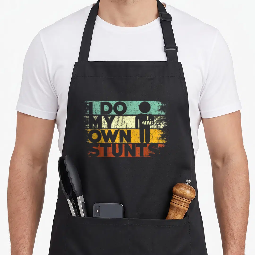 Stunt recovery day apron - I DO MY OWN STUNTS text over teal yellow red grey bands with stick figure — shown on a black cotton canvas apron against a white background