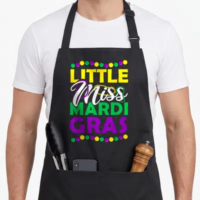 Miss mardi gras aprons - LITTLE Miss MARDI GRAS text with purple green yellow beads stacked vertically — shown on a black cotton canvas apron against a white background