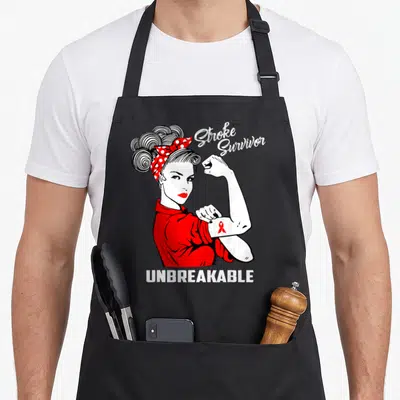 Survivor stroke awareness apron - Cartoon woman flexing arm with red ribbon and text Stroke Survivor Unbreakable — shown on a black cotton canvas apron against a white background