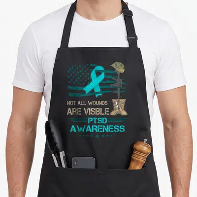 Ptsd patriotic apron - Distressed military memorial with teal ribbon, helmet, rifle, boots, and PTSD Awareness text — shown on a black cotton canvas apron against a white background