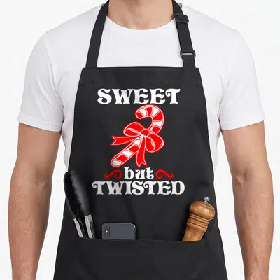 Sweet candy cane apron - SWEET but TWISTED text with red and white striped candy cane and bow — shown on a black cotton canvas apron against a white background
