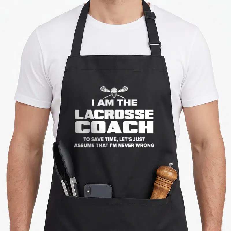 Lacrosse appreciation apron - White lacrosse helmet and crossed sticks above bold text reading I am the lacrosse coach — shown on a black cotton canvas apron against a white background