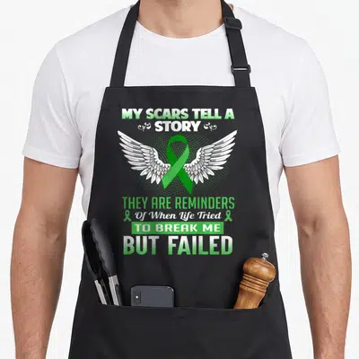 Kidney story of the apron - Green ribbon with white wings and MY SCARS TELL A STORY text — shown on a black cotton canvas apron against a white background