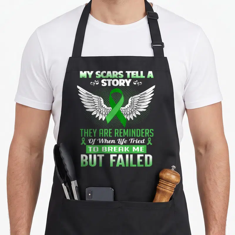 Kidney story of the apron - Green ribbon with white wings and MY SCARS TELL A STORY text — shown on a black cotton canvas apron against a white background