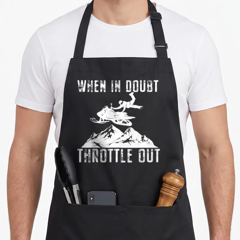 Sled winter apron - White distressed text When In Doubt Throttle Out with snowmobile and mountain silhouette — shown on a black cotton canvas apron against a white background