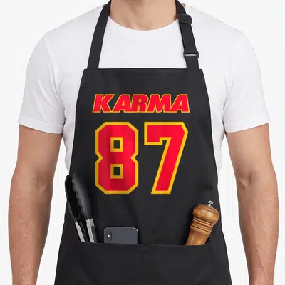 Cute karma apron - Red and yellow text reading KARMA 87 stacked vertically on a dark background — shown on a black cotton canvas apron against a white background