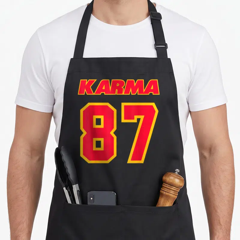 Cute karma apron - Red and yellow text reading KARMA 87 stacked vertically on a dark background — shown on a black cotton canvas apron against a white background