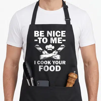Retro culinary aprons - White distressed text BE NICE TO ME I COOK YOUR FOOD above crossed cleavers and chef hat — shown on a black cotton canvas apron against a white background