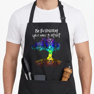 Chakra tree apron - Rainbow tree with seven chakra symbols and white text Be the ENERGY you WANT to attract — shown on a black cotton canvas apron against a white background