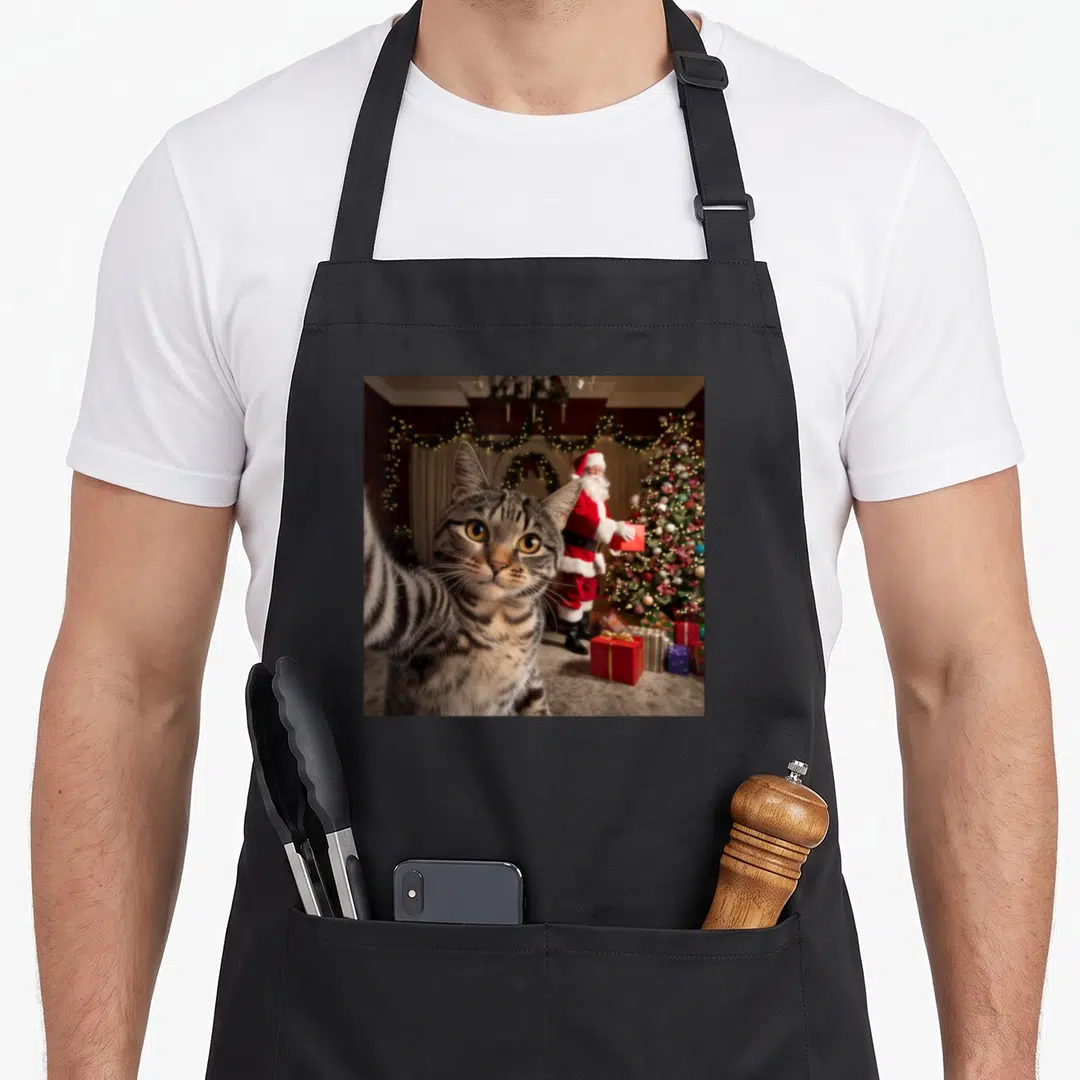 Christmas funny cat apron - Tabby cat selfie pose with Santa Claus and Christmas tree in red and gold — shown on a black cotton canvas apron against a white background