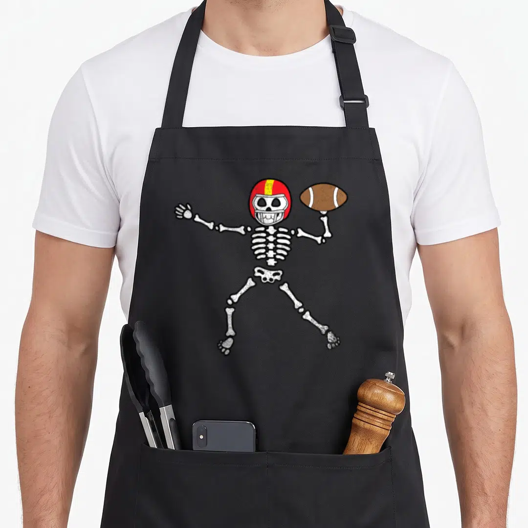 Skeleton football halloween apron - Cartoon white skeleton in red and yellow helmet holding brown football with arm — shown on a black cotton canvas apron against a white background