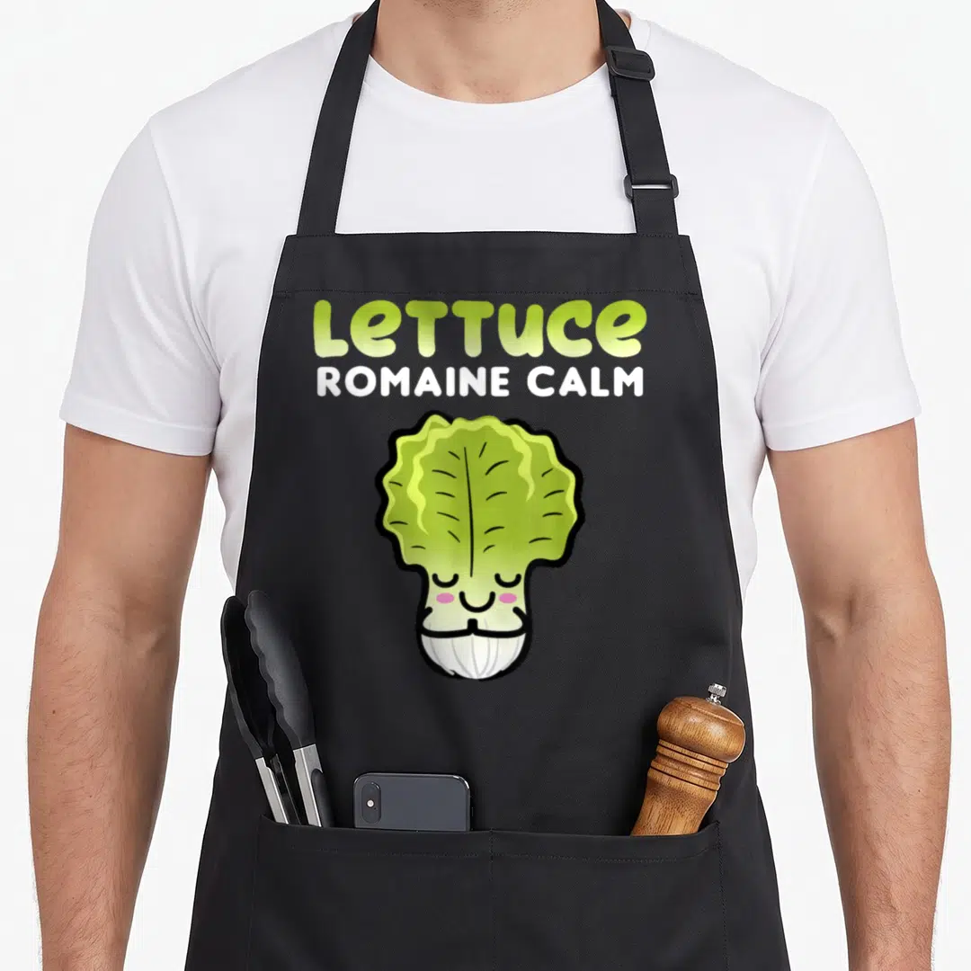 Based veggie apron - Cartoon lettuce with pink cheeks and text LETTUCE ROMAINE CALM in lime and white — shown on a black cotton canvas apron against a white background