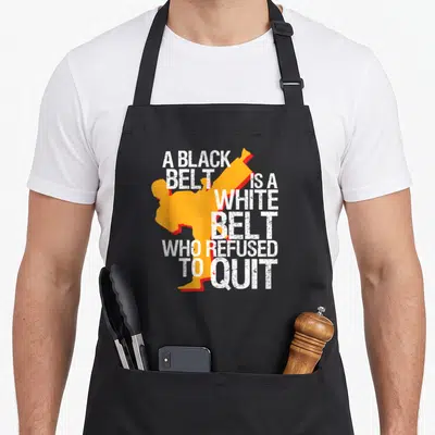 Taekwondo student aprons - Distressed white text A BLACK BELT IS A WHITE BELT WHO REFUSED TO QUIT wraps around — shown on a black cotton canvas apron against a white background
