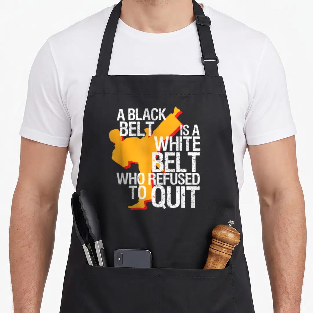 Taekwondo student aprons - Distressed white text A BLACK BELT IS A WHITE BELT WHO REFUSED TO QUIT wraps around — shown on a black cotton canvas apron against a white background