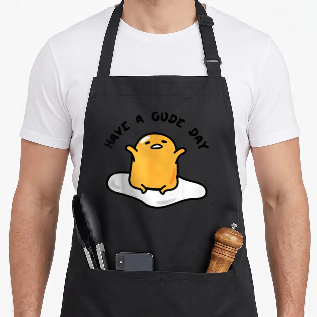 Gude gudetama apron - Orange egg yolk character on white base with arched HAVE A GUDE DAY text — shown on a black cotton canvas apron against a white background