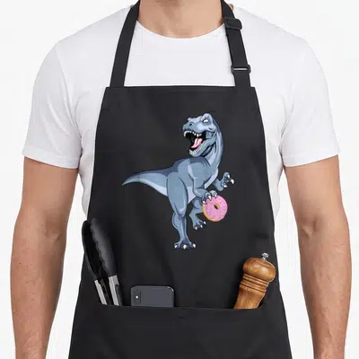 Dinosaur christmas apron - Cartoon blue-grey T-Rex holding a pink frosted donut with sprinkles — shown on a black cotton canvas apron against a white background
