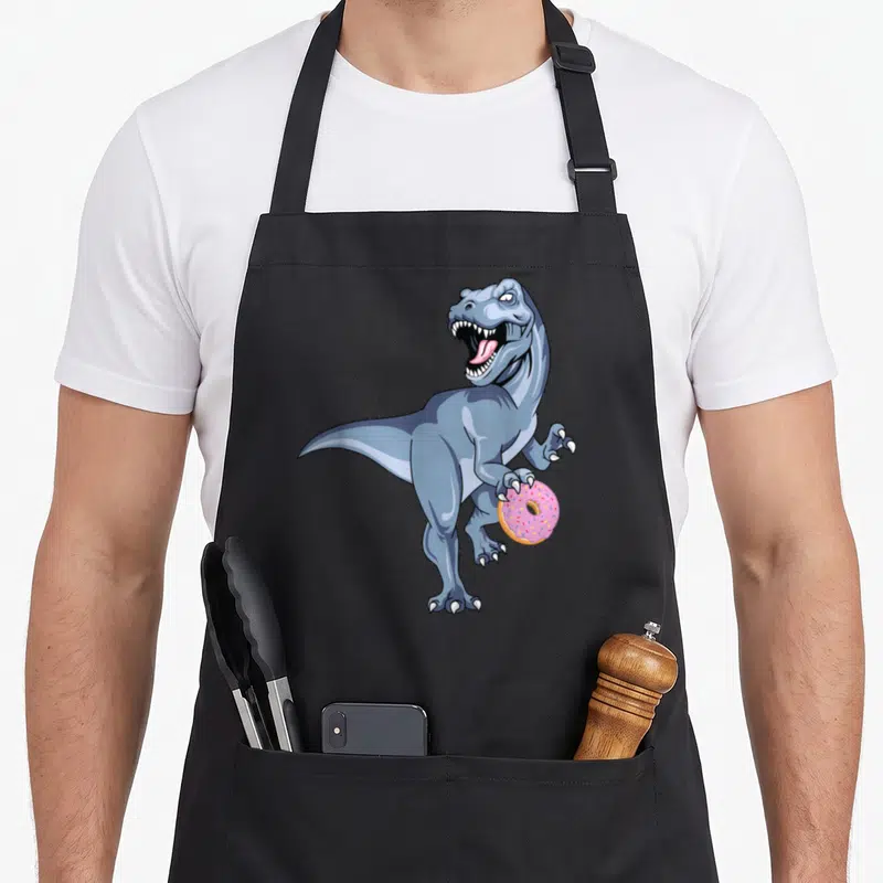 Dinosaur christmas apron - Cartoon blue-grey T-Rex holding a pink frosted donut with sprinkles — shown on a black cotton canvas apron against a white background