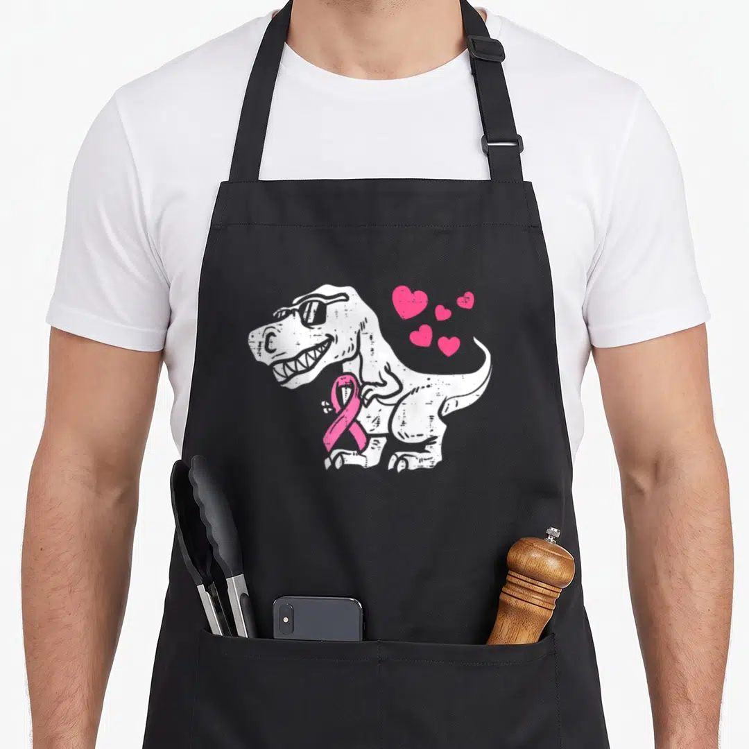 Ribbon breast cancer awareness aprons - White T-Rex with sunglasses holding pink ribbon and floating hearts — shown on a black cotton canvas apron against a white background