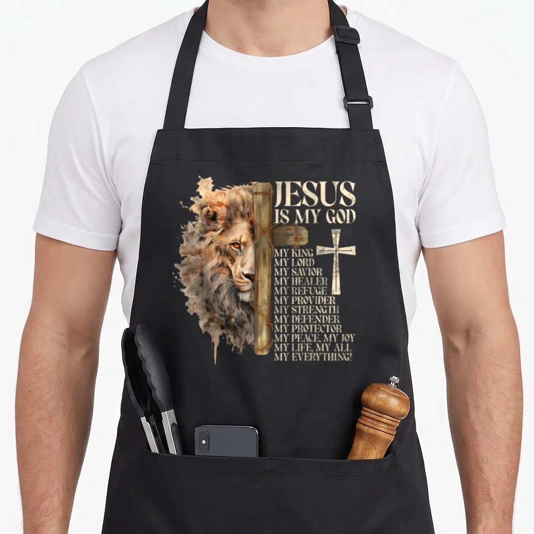 Faith lions apron - Watercolor lion head behind wooden cross with stacked JESUS IS MY GOD text in beige and brown — shown on a black cotton canvas apron against a white background