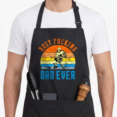 Pucking father day apron - Retro hockey player illustration with orange blue yellow red striped sun and text — shown on a black cotton canvas apron against a white background