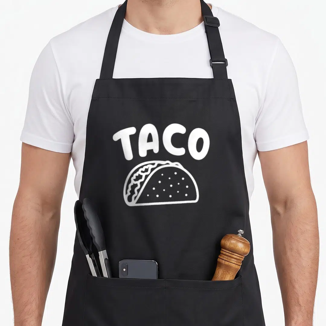 Taquito mexican apron - White TACO text arched over a cartoon taco outline on black fabric — shown on a black cotton canvas apron against a white background