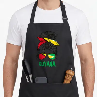 Funny guyanese guyana apron - Messy bun hair with red yellow green bandana and flag sunglasses above green GUYANA text — shown on a black cotton canvas apron against a white background